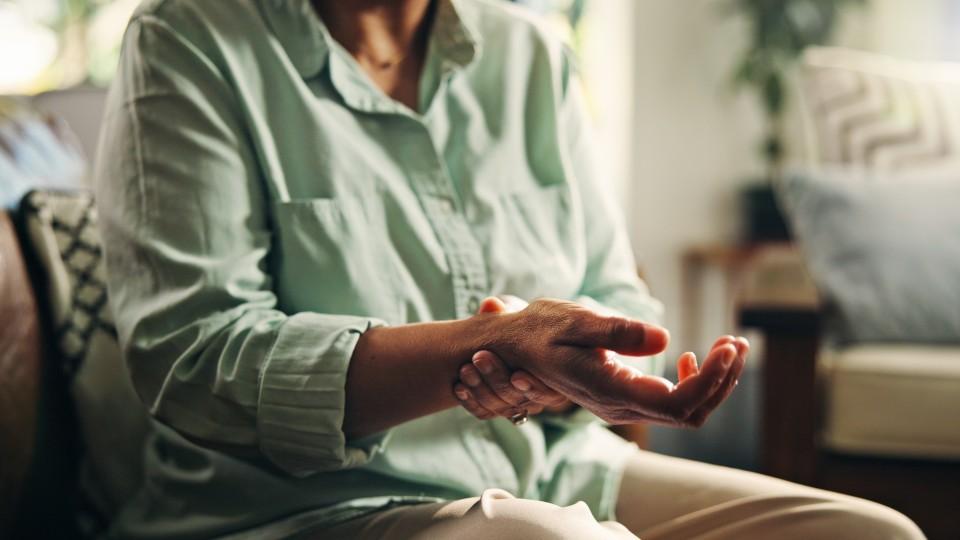 An older woman with rheumatoid arthritis holds her wrist in pain while sat in an armchair