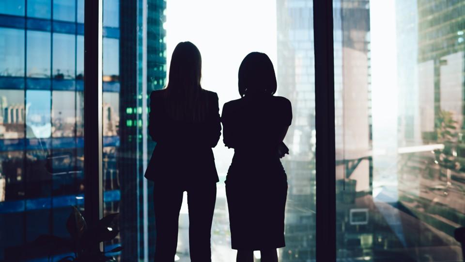 Two female silhouettes in a city office overlook the urban landscape below
