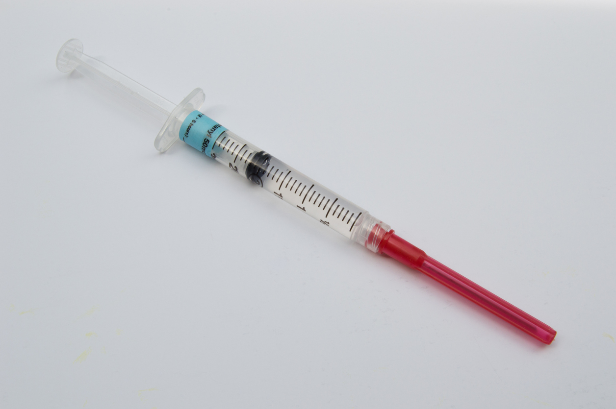 A syringe containing the pain medicine, fentanyl, topped with a red capped safety needle and photographed against a white background from above.