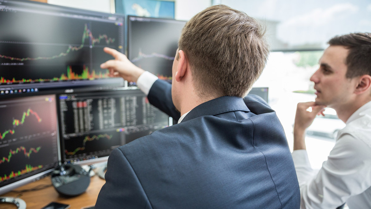 Businessmen trading stocks online. Stock brokers looking at graphs, indexes and numbers on multiple computer screens. Colleagues in discussion in traders office. Business success concept.