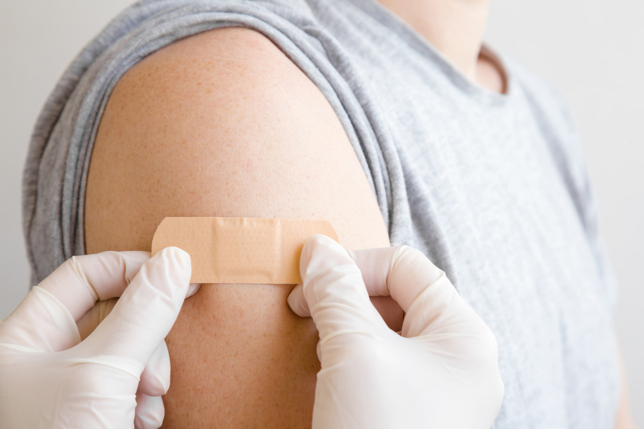 Doctor in white rubber protective gloves putting adhesive bandage on young man's arm after scratch on skin or injection of vaccine. First aid. Medical, pharmacy and healthcare concept. Closeup.