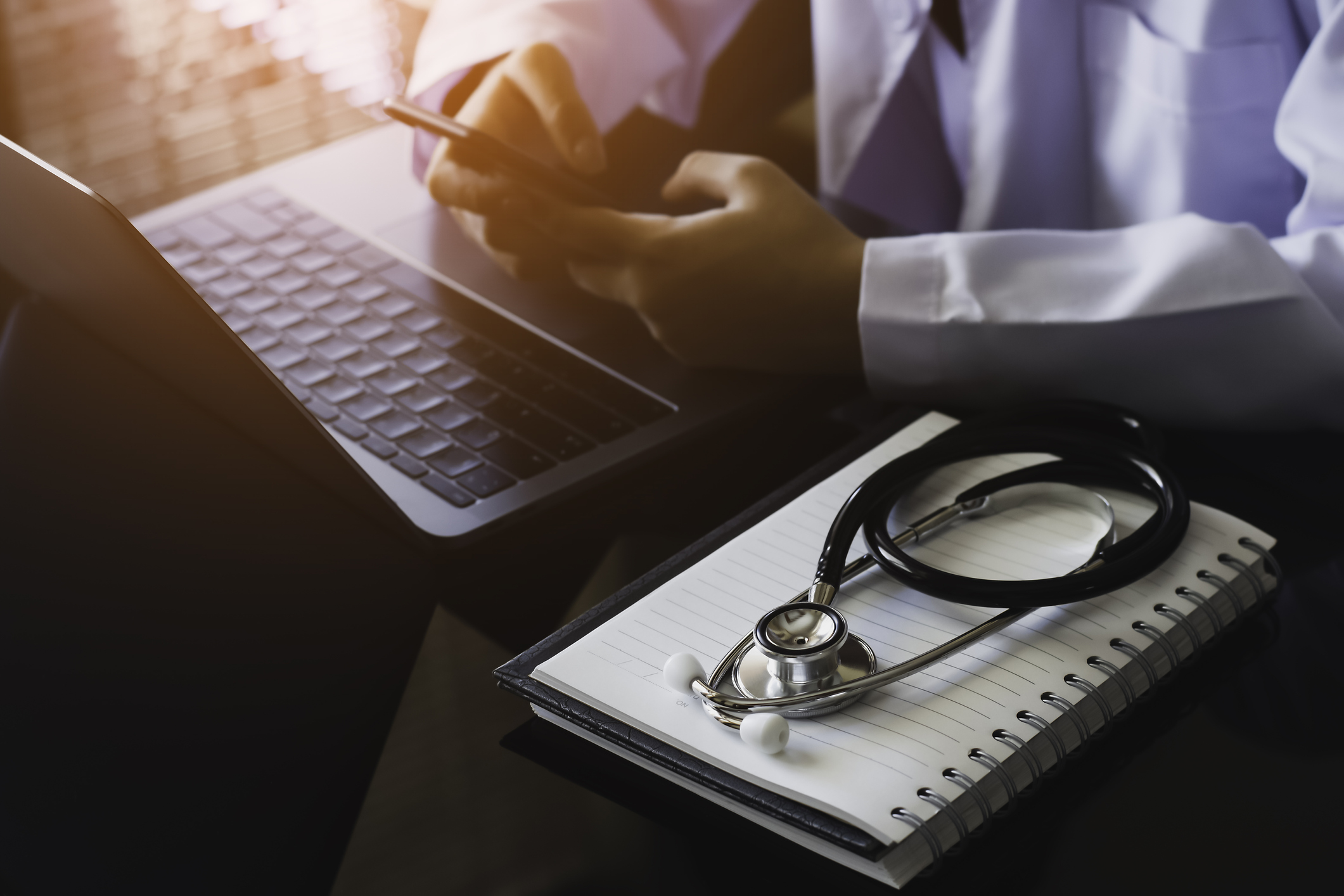 Doctor using mobile smart phone, work on laptop computer with notebook and medical stethoscope on the desk at office. Telehealth, telemedicine or medic tech concept.