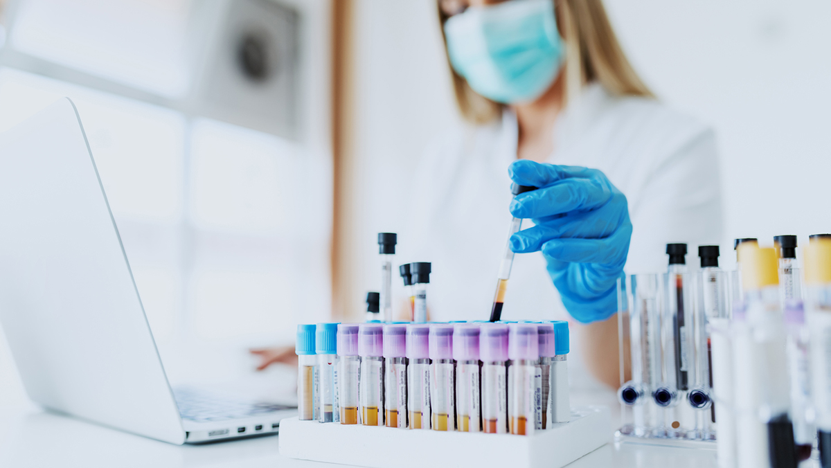 Close up of lab assistant in uniform, with mask and rubber gloves holding test tube with blood sample while sitting on chair and typing on laptop. Selective focus on test tubes.